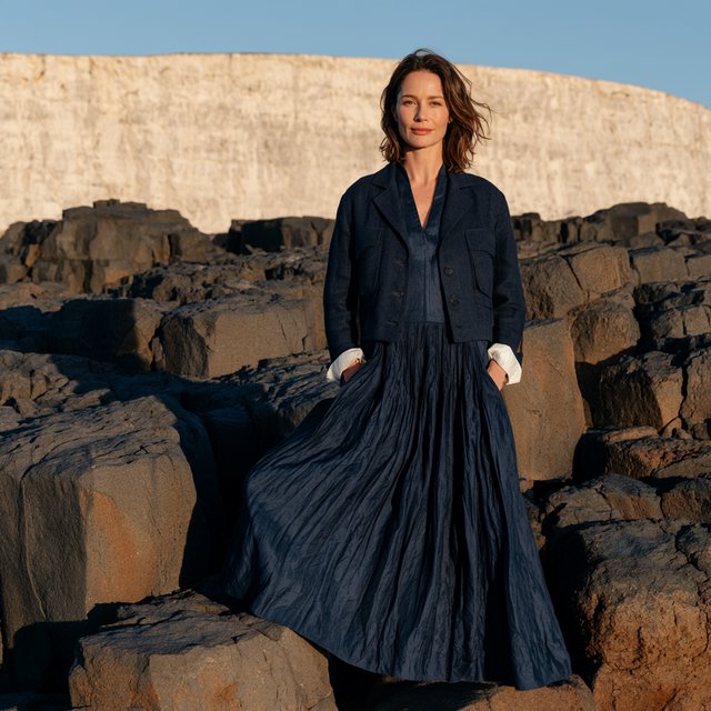 Across a basalt terrace, an adult woman in her early forties poses in a full-length fashion photograph, wearing a pleated indigo silk dress and cropped linen jacket, her composed half-smile lit by hard afternoon sunlight from one side against a pale limestone ridge. — Midjourney V8.2