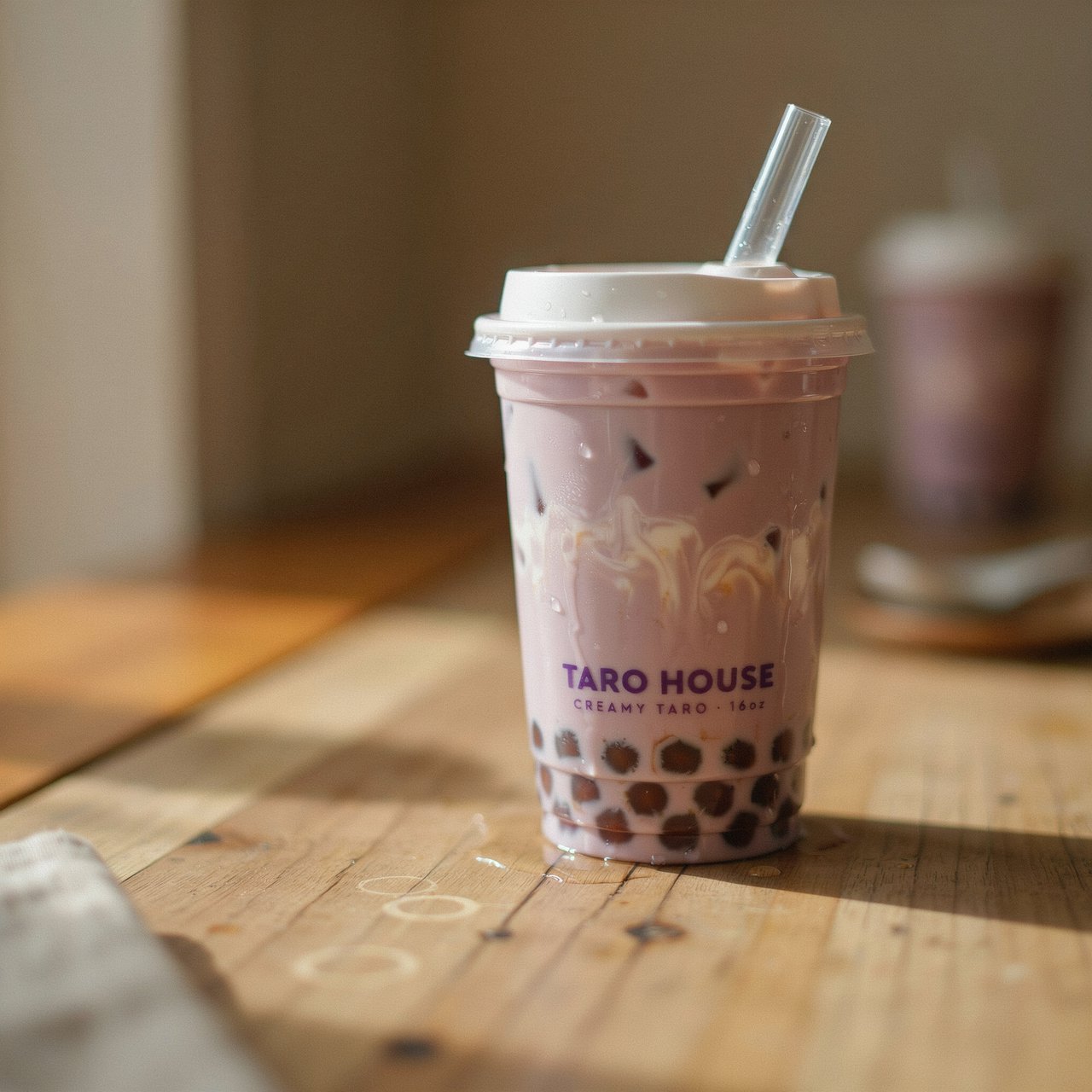 Photorealistic product shot of iced taro bubble tea in a clear frosted plastic cup, condensation beading down curved sides, wide clear straw piercing matte white domed lid. Plump dark brown tapioca pearls settled at the base, swirled creamy purple tea with soft marbled cream streaks, fine ice crystals glinting inside. Resting on weathered light oak table, soft golden hour side lighting casting gentle long shadows, warm neutral beige background. Shallow depth of field, sharp focus on glass texture and pearl details, professional food photography aesthetic, soft moody warm colour palette with rich lavender and caramel tones. — Ideogram v4
