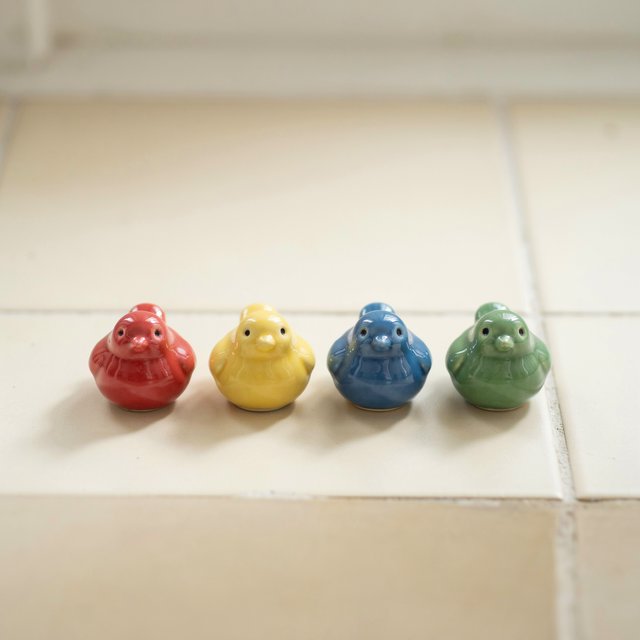 On a sunlit tiled ledge, exactly four ceramic bird figurines form a row: red at far left, yellow at left-center, blue at right-center, and green at far right; all face forward, nothing else. — Ideogram v4