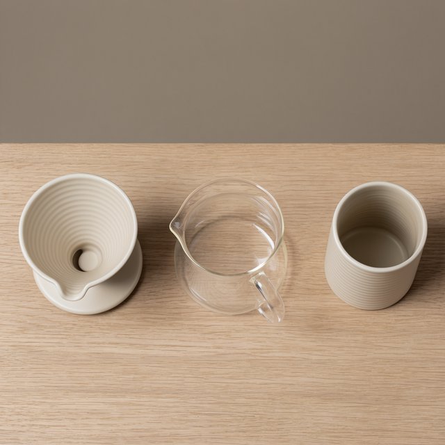 From directly overhead, an unbranded pour-over coffee set with ribbed porcelain and clear borosilicate components arranged in sequence on pale oak; warm taupe background, broad diffused softbox light and gentle side fill shape this commercial product image, no lettering. — Model 11