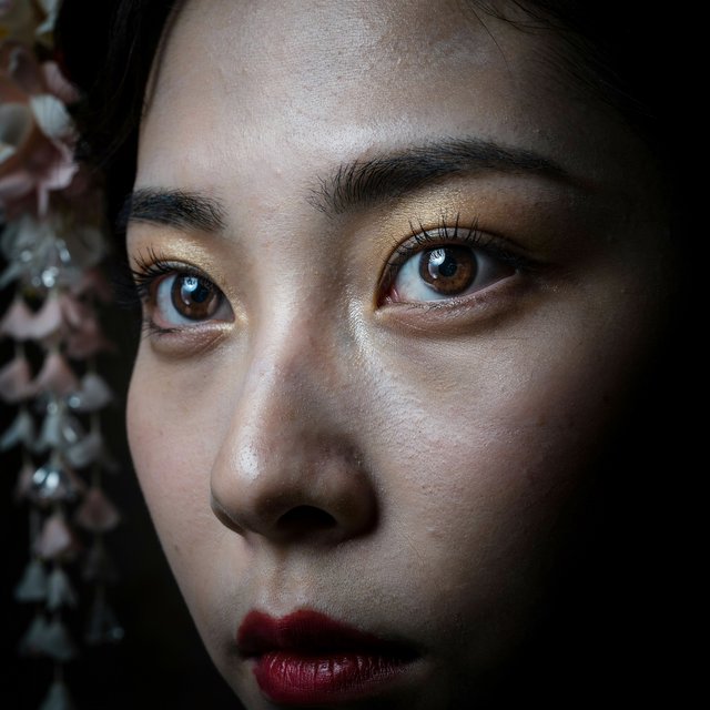 Extreme close-up macro photography of a Geisha's face. The focus is on her eyes, revealing intense emotion and hyper-realistic detail. Her skin is covered in traditional white 'Oshiroi' makeup, showing subtle, realistic cracking and skin texture. Dramatic Chiaroscuro lighting with deep shadows and a single soft light source highlighting the shimmer in her golden eyeshadow and the wetness of her eyes. Ornate kanzashi (hair ornaments) visible in the blurred periphery with intricate silk flowers and dangling silver petals. Cinematic composition, shot on 100mm macro lens, incredibly detailed eyelashes and iris patterns. — Model 8