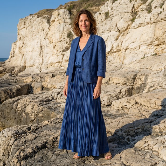 Across a basalt terrace, an adult woman in her early forties poses in a full-length fashion photograph, wearing a pleated indigo silk dress and cropped linen jacket, her composed half-smile lit by hard afternoon sunlight from one side against a pale limestone ridge. — Qwen Image 3.0 Pro