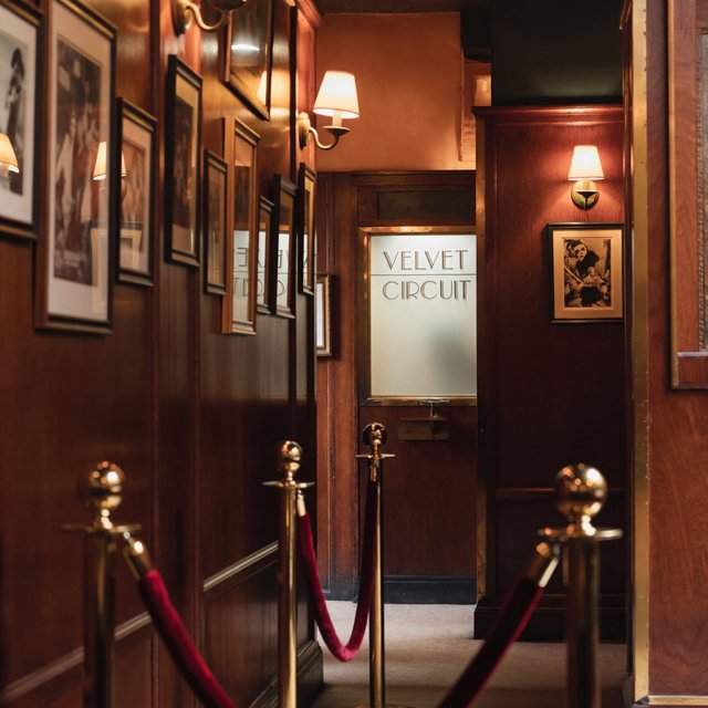 A narrow neighborhood cinema lobby glows with amber sconces, velvet ropes, and framed film stills; "VELVET CIRCUIT" is etched in elegant silver lettering across the frosted glass ticket-booth window. — ImagineArt 2