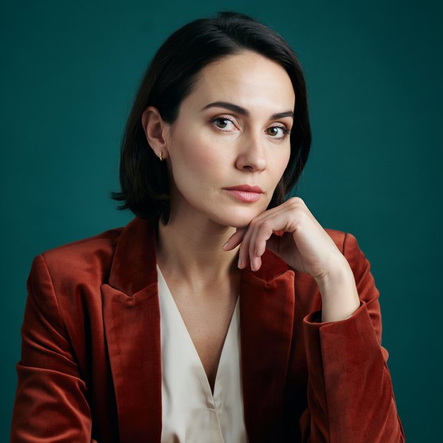 Portrait of an adult woman in her 30s wearing a rust-colored velvet jacket over an ivory blouse, one hand resting beneath her chin, thoughtful expression, deep teal studio backdrop, soft side lighting and gentle cheek highlights. — Ideogram v4