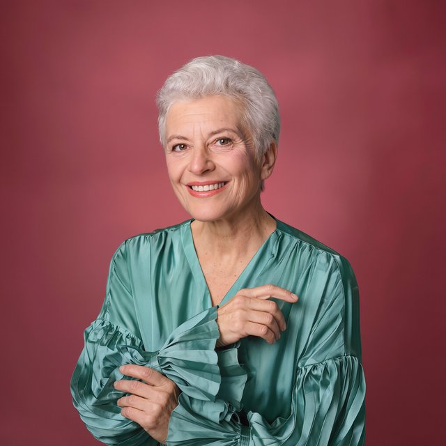 Portrait photograph of an adult woman in her early 60s wearing a softly pleated sea-green silk blouse, hands brushing its gathered sleeve fabric. Her amused, open expression stands against a matte rose backdrop, illuminated by broad, gentle studio light. — Kling v3