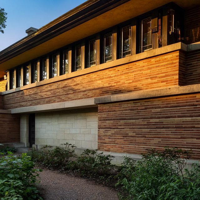 Exterior of a Prairie School residence with broad overhanging eaves, warm brick, limestone courses, and ribbon windows; amber evening light grazes the facade, viewed obliquely from a garden path, with disciplined horizontal lines and credible structure. — Krea 2 Large