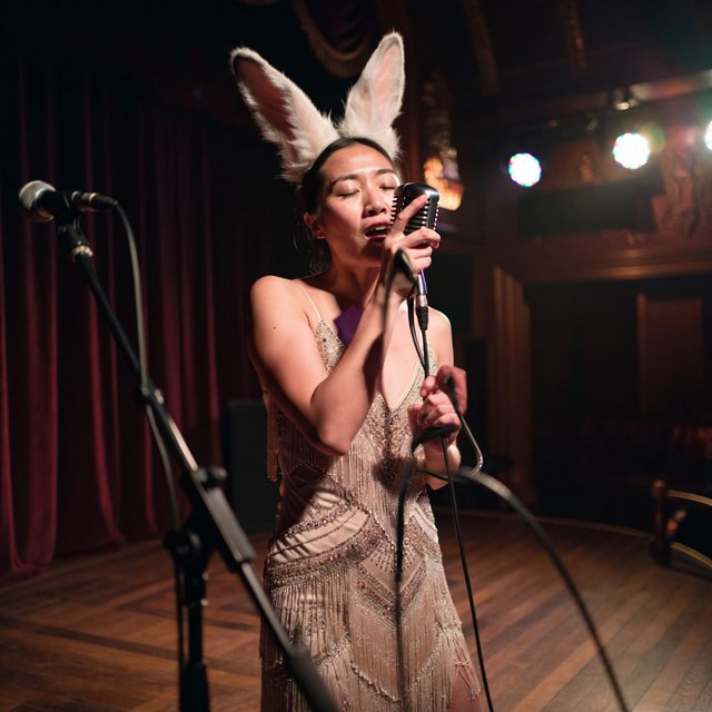 Anthropomorphic rabbit-human hybrid. No human ears visible; the areas where ears would normally be are covered by rabbit fur. A pulchritudinous East Asian 32-year-old wearing a luxurious flapper dress with a deep V split down the center, standing on stage behind a luxurious retro microphone, singing her heart out into it. Taken with a professional DSLR camera and a 35mm lens. Background bokeh, stage lights with a bloom effect. — ImagineArt 2