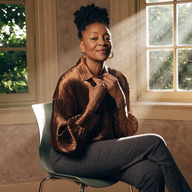 Portrait of an adult Black woman in her 50s wearing a pleated copper satin blouse and tailored charcoal trousers, her hands gently holding the blouse fabric, calm half-smile, warm plaster backdrop, late-afternoon window light grazing her face. — Recraft v4