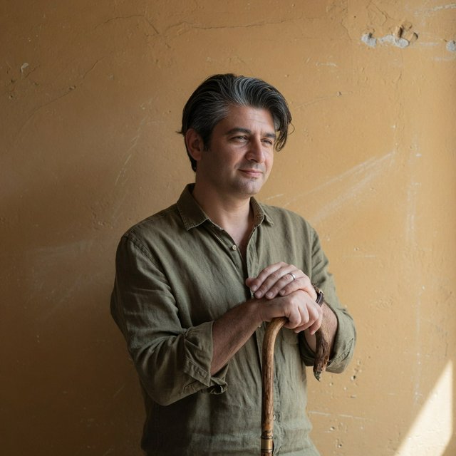 Portrait photograph of an adult man in his 40s with silver-streaked hair, wearing a textured olive linen shirt and rolled sleeves, hands resting on a wooden cane, relaxed expression, ochre plaster backdrop, soft window light from the side. — Grok Imagine Image Quality