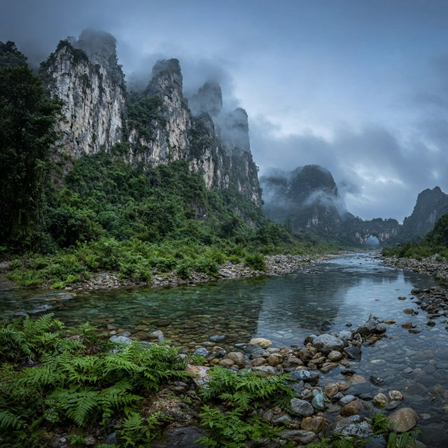 A limestone karst valley at blue hour after a light shower, towering pale cliffs rising above a clear river, low clouds snagging on pinnacles, slick ferns and rounded pebbles leading toward a distant stone arch. — Grok Imagine Image 2