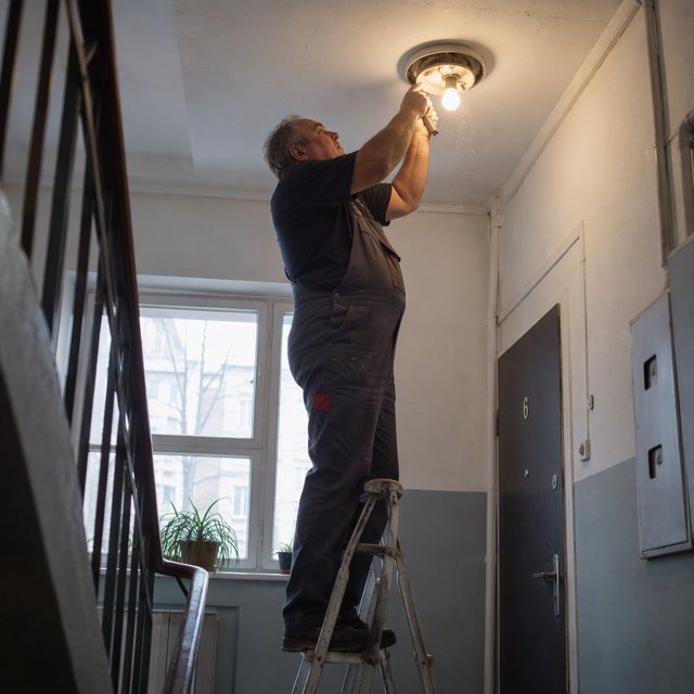 Candid documentary photograph of an adult building caretaker changing a ceiling light in a quiet apartment stairwell, pale afternoon window light, 35mm lens with moderate depth of field, plaster dust drifting from the fixture. — GPT Image 2.5 Sunburst