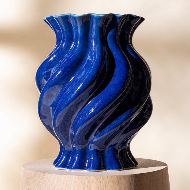 Commercial product photograph of an unbranded sculptural ceramic vase, formed from glossy cobalt porcelain, standing on a pale oak pedestal against a warm ivory background, illuminated by soft directional daylight and subtle fill. — Recraft v4 Pro