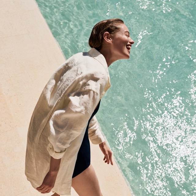 Beside a pale-stone pool, an adult woman in a navy one-piece swimsuit and loose ivory linen shirt pauses mid-laugh, sunlight sparkling on water and warm stone; crisp shadows and poised styling give the moment polished fashion-magazine glamour. — Midjourney V8.2