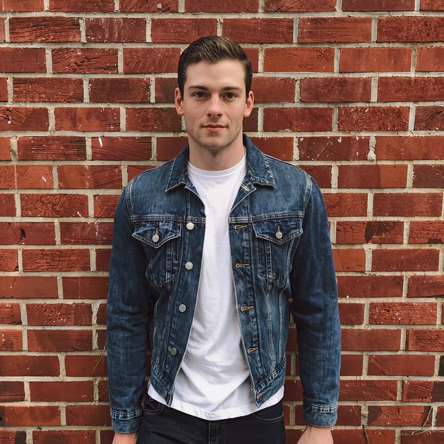 Environmental portrait of a young man in a denim jacket leaning on a red brick wall, overcast light, looking just past the camera. — Kling v3