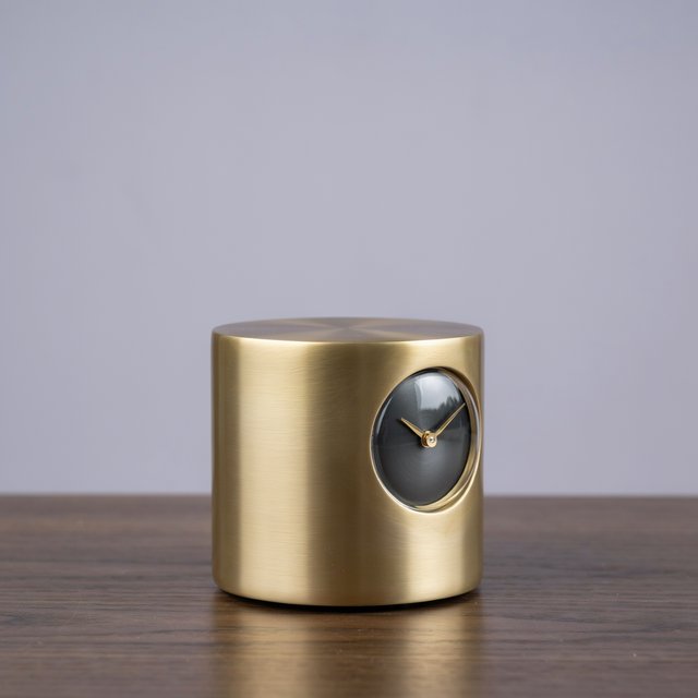 Commercial product photograph of an unbranded sculptural table clock made from satin brass and smoked glass, resting on a dark walnut surface against a muted lavender background, illuminated by a large diffused softbox from the upper left and a subtle rear rim light. — Ideogram v4