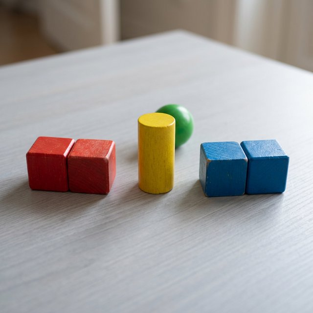 Exactly five wooden toy blocks sit on a pale gray table: two red cubes on the left, one yellow cylinder in the center, and two blue cubes on the right. A small green ball sits behind the cylinder, nothing else. — Nano Banana 2.1