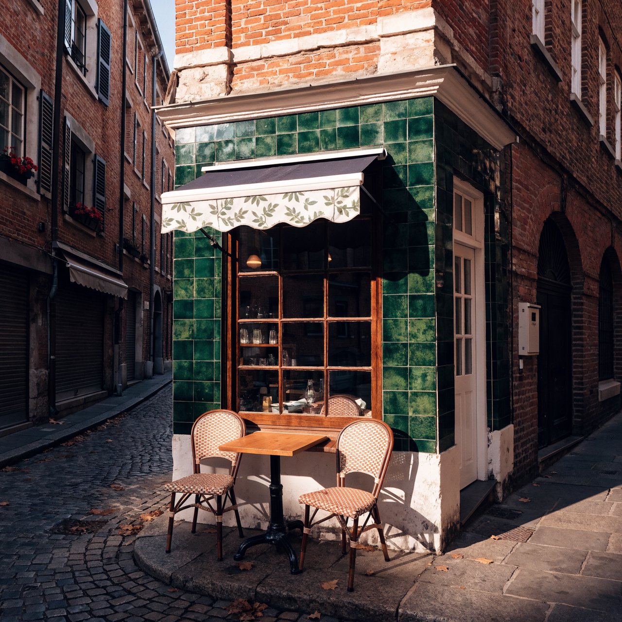 A tiny corner café in a narrow European street, green tiled facade, two bistro tables, morning light, no people. — Recraft v4 Pro