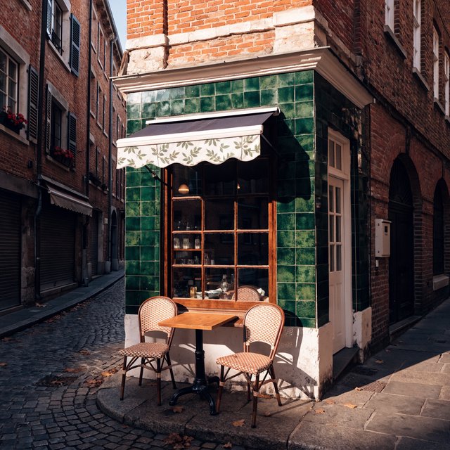A tiny corner café in a narrow European street, green tiled facade, two bistro tables, morning light, no people. — Recraft v4 Pro