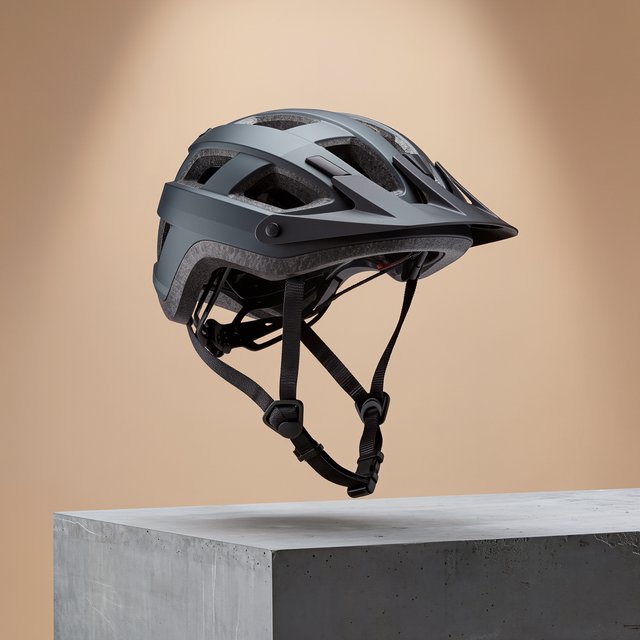 Commercial product photograph of an unbranded folding bicycle helmet made from matte charcoal polymer with soft gray padding, resting on a honed concrete plinth against a muted sand background, illuminated by a broad overhead softbox and gentle rim light. — Kling v3