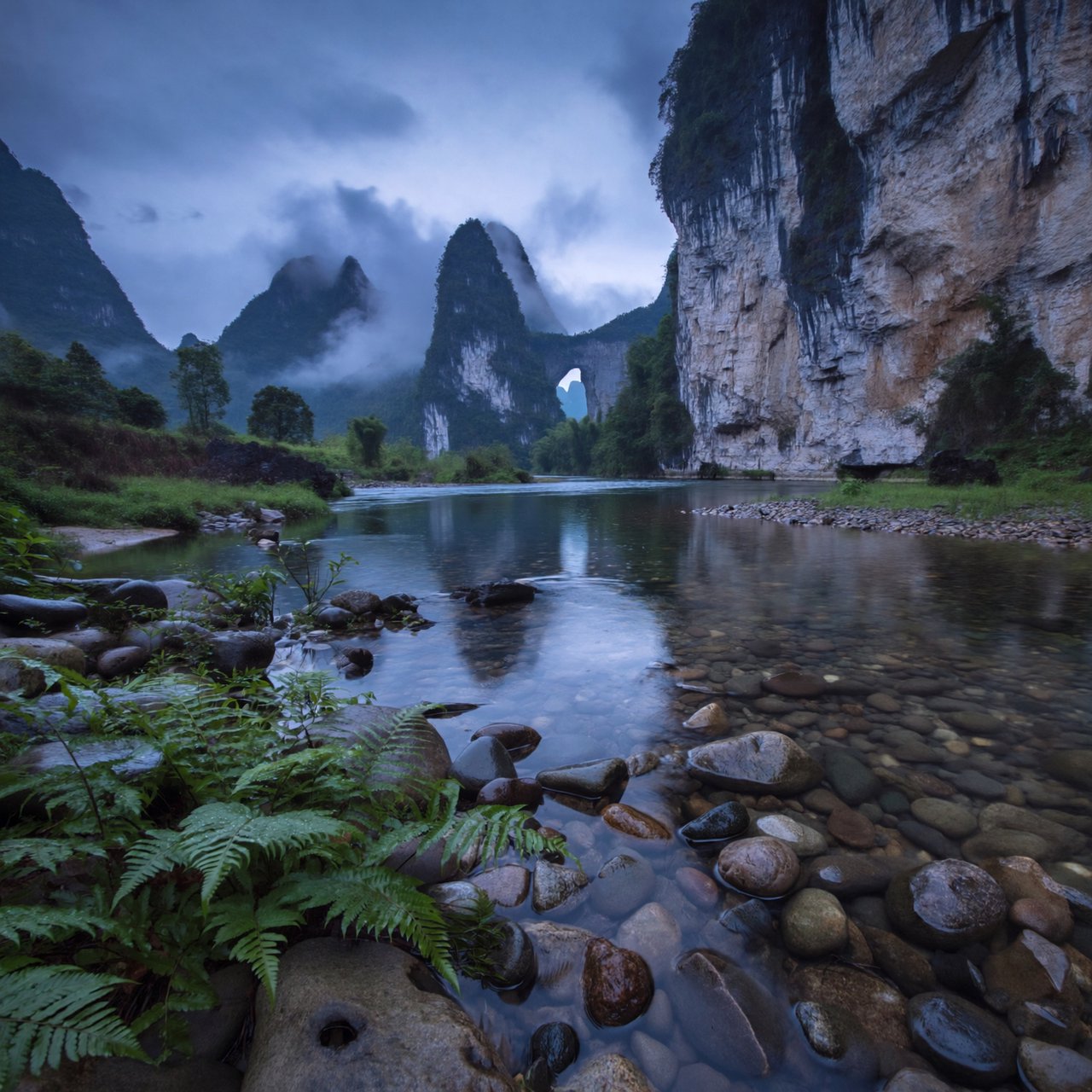 A limestone karst valley at blue hour after a light shower, towering pale cliffs rising above a clear river, low clouds snagging on pinnacles, slick ferns and rounded pebbles leading toward a distant stone arch. — ImagineArt 2
