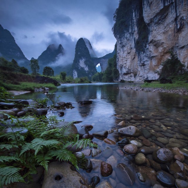 A limestone karst valley at blue hour after a light shower, towering pale cliffs rising above a clear river, low clouds snagging on pinnacles, slick ferns and rounded pebbles leading toward a distant stone arch. — ImagineArt 2