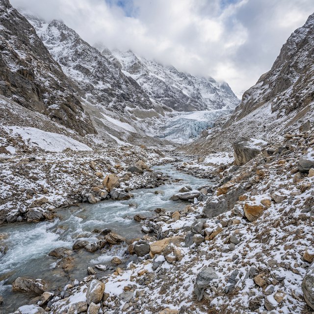 Beyond a braided glacier-fed stream, a steep moraine valley opens beneath clearing dawn snow flurries; gritty ice grains and water-polished stones sharpen the near bank, while layered ridges fade into cloud. — Model 18