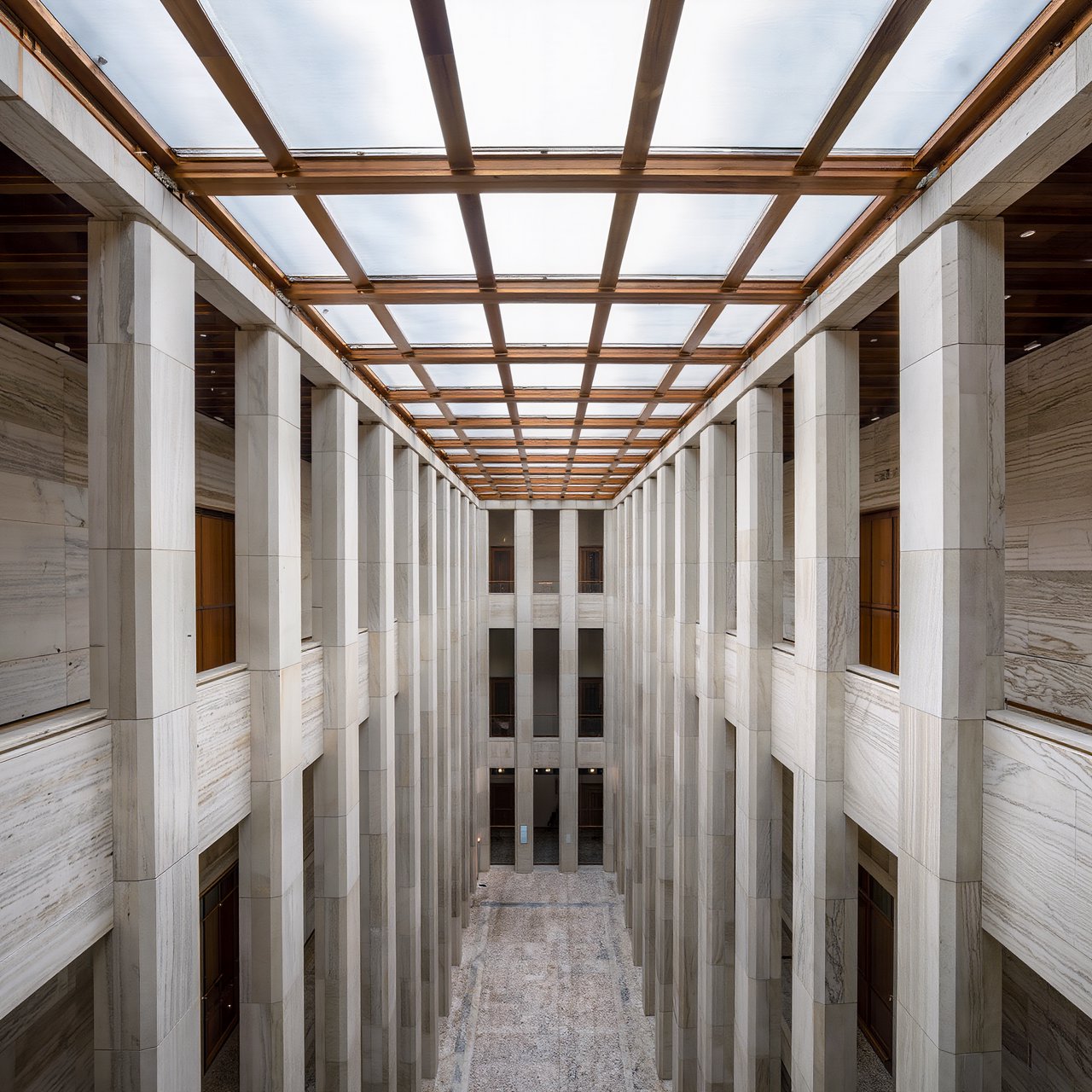 A soaring civic atrium in contemporary Italian Rationalist style pairs pale travertine, bronze mullions, and terrazzo beneath a glazed roof; cool morning light falls from above, viewed from a mezzanine, with aligned bays and credible supports. — Kling v3
