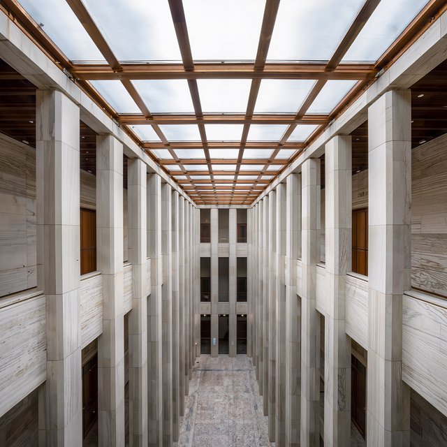 A soaring civic atrium in contemporary Italian Rationalist style pairs pale travertine, bronze mullions, and terrazzo beneath a glazed roof; cool morning light falls from above, viewed from a mezzanine, with aligned bays and credible supports. — Kling v3