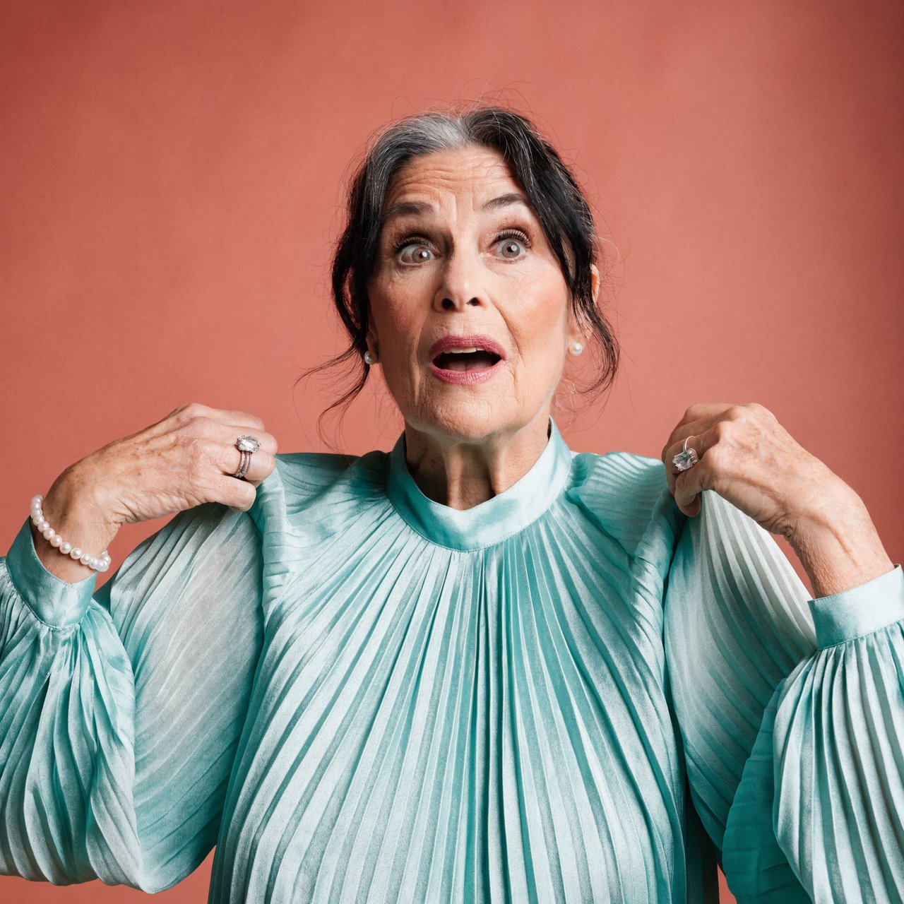 Portrait photograph of an adult woman in her early 60s wearing a softly pleated sea-green silk blouse, hands brushing its gathered sleeve fabric. Her amused, open expression stands against a matte rose backdrop, illuminated by broad, gentle studio light. — Recraft v4 Pro