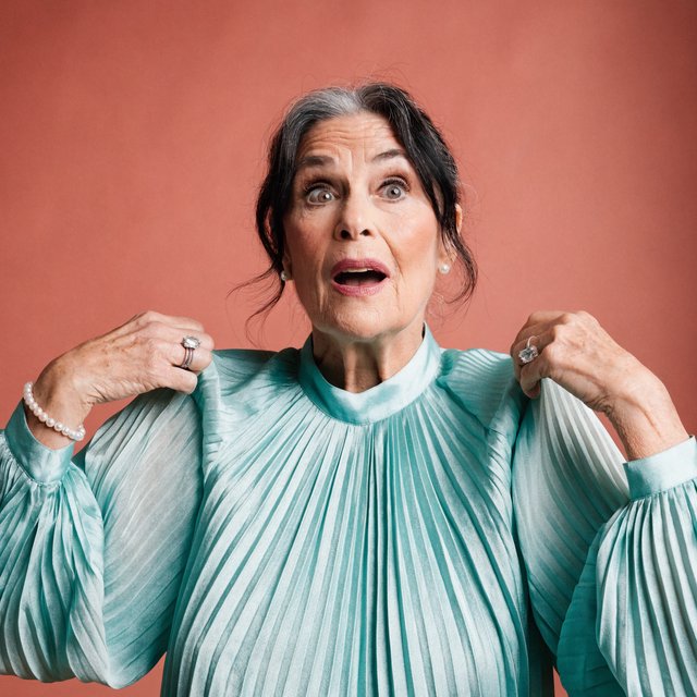 Portrait photograph of an adult woman in her early 60s wearing a softly pleated sea-green silk blouse, hands brushing its gathered sleeve fabric. Her amused, open expression stands against a matte rose backdrop, illuminated by broad, gentle studio light. — Recraft v4 Pro