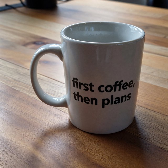 A white ceramic mug on a desk with the words "first coffee, then plans" printed in black lowercase sans-serif. — ImagineArt 1.5 Pro