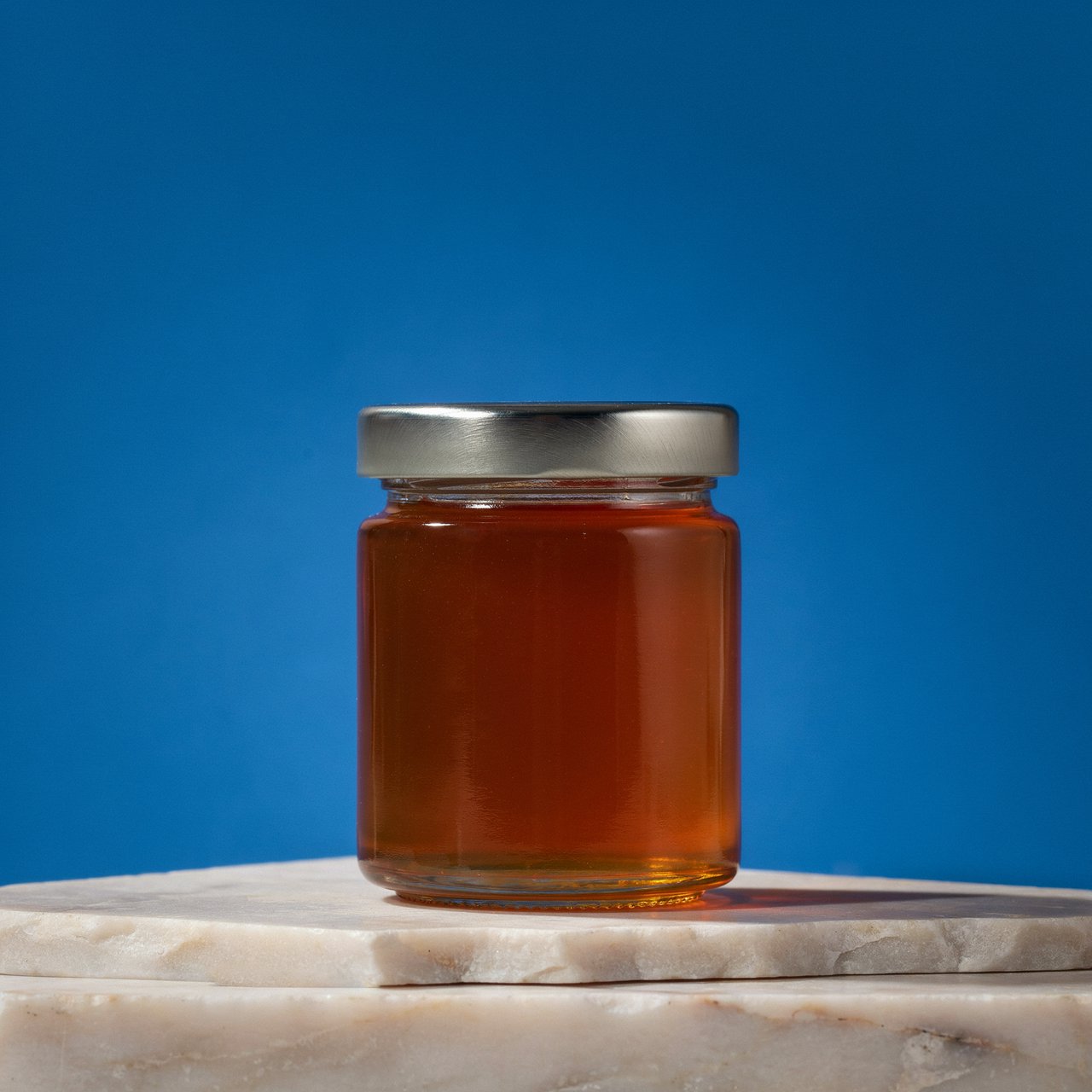 A squat unbranded honey jar with clear glass walls and a plain brushed-aluminum lid rests on honed pale limestone; broad diffused key light and warm rim light shape the commercial product image against a seamless cobalt-blue background, with no lettering. — Ideogram v4