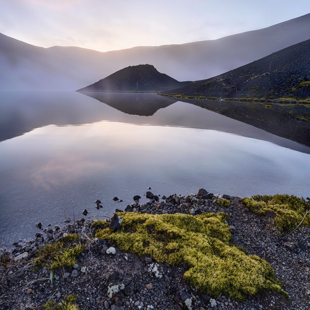At first light, a rain-cleared volcanic caldera lake lies beneath thinning fog; pumice grains and damp lichen sharpen the near shore, while dark cinder slopes step back toward a distant rim. — Kling v3