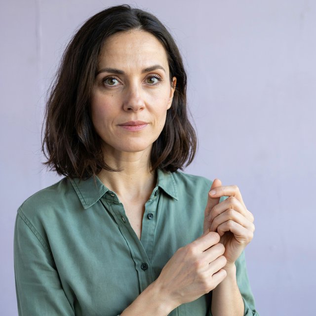 Portrait photograph of an adult woman in her late 30s wearing a moss-green cotton poplin shirt, fingers lightly turning its rolled cuff. Her calm, observant expression faces the camera against a chalky lavender backdrop, lit by soft diffused skylight. — Ideogram v4.5