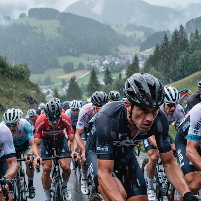A frontal, close-up shot of a field of competitive cyclists ascending a mountain in the rain. In the background, we see a lush green valley with a small town. — ImagineArt 2