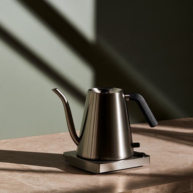Morning daylight spills across a kitchen counter, revealing an unbranded electric kettle in satin stainless steel with a ribbed heat-resistant polymer handle. It sits on honed limestone against a muted sage backsplash, with window light and white bounce fill; commercial product image, no lettering. — Midjourney V8.2