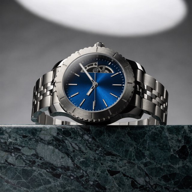Commercial product photograph of an unbranded mechanical wristwatch with a brushed titanium case and deep blue enamel dial, resting on dark green marble against a soft gray background, illuminated by a large diffused softbox and narrow edge light. — Recraft v4 Pro
