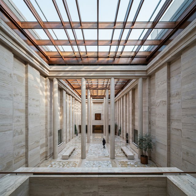 A soaring civic atrium in contemporary Italian Rationalist style pairs pale travertine, bronze mullions, and terrazzo beneath a glazed roof; cool morning light falls from above, viewed from a mezzanine, with aligned bays and credible supports. — Ideogram v4