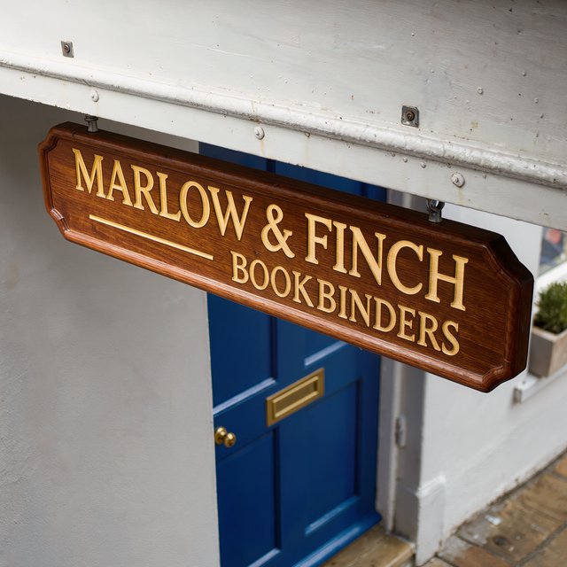 A hanging wooden shop sign that reads "MARLOW & FINCH — BOOKBINDERS" in gold serif letters, carved, above a blue door. — Kling v3