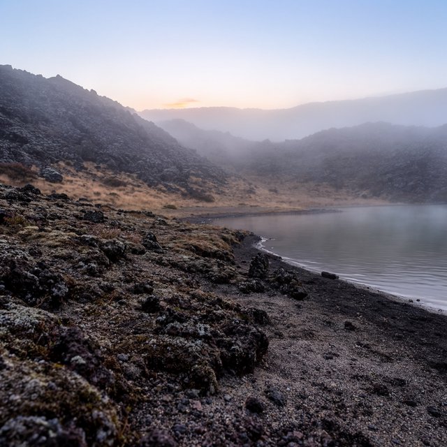 At first light, a rain-cleared volcanic caldera lake lies beneath thinning fog; pumice grains and damp lichen sharpen the near shore, while dark cinder slopes step back toward a distant rim. — Krea 2 Large