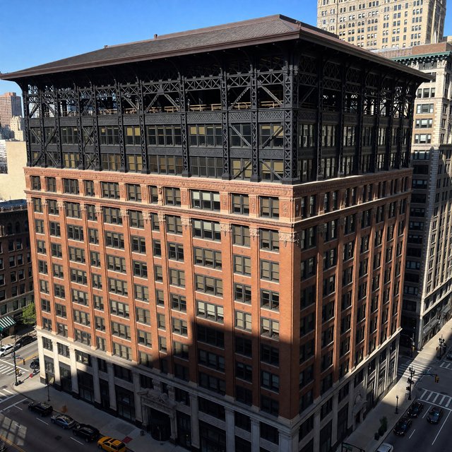 Exterior of a Chicago School office tower, with a visible steel-frame grid, warm terra-cotta cladding, broad rectangular windows, and a flat roofline; clear afternoon light, viewed from across a city street, with precise verticals and believable construction. — ImagineArt 1.5 Pro
