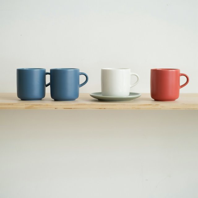Exactly four ceramic mugs sit on a pale wooden shelf: two blue mugs on the left, one white mug in the center, and one red mug on the right. A green saucer is beneath the center mug, nothing else. — Ideogram v4