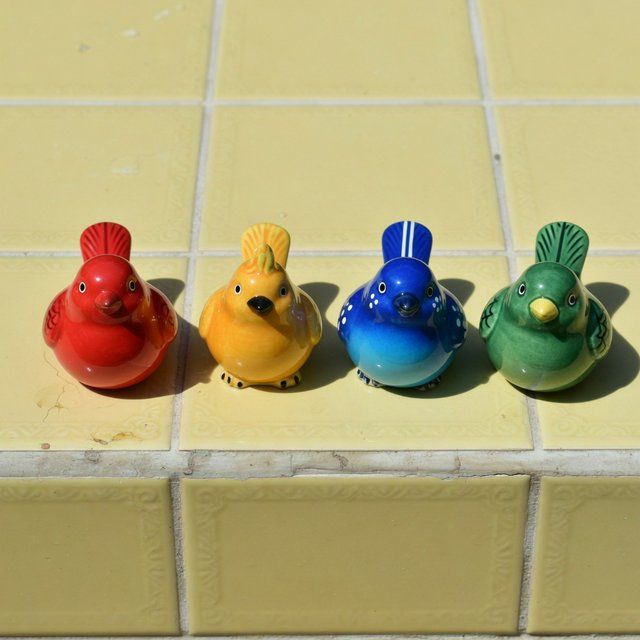 On a sunlit tiled ledge, exactly four ceramic bird figurines form a row: red at far left, yellow at left-center, blue at right-center, and green at far right; all face forward, nothing else. — Grok Imagine Image Quality