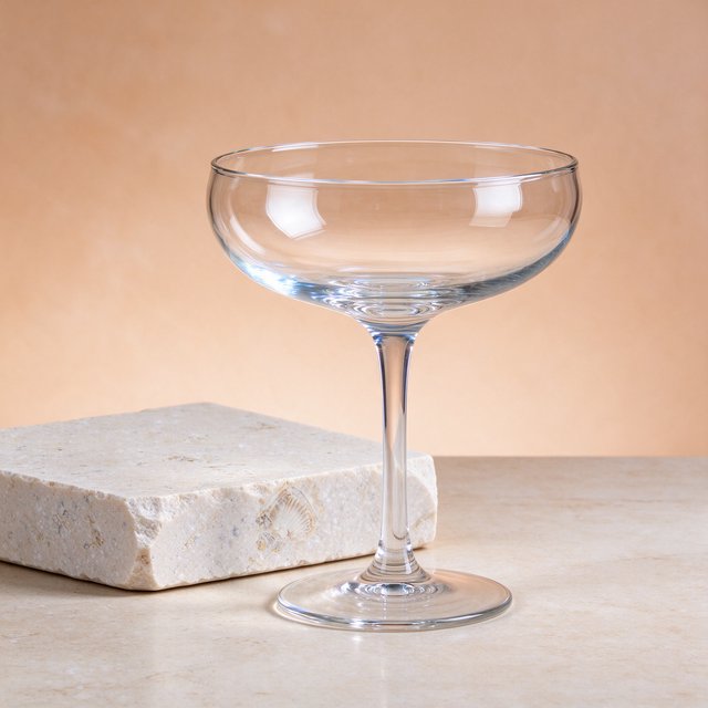 Beside a pale limestone block, an unbranded handblown cocktail coupe in clear soda-lime glass catches crisp window-shaped highlights; its curved bowl bends a muted apricot backdrop under broad diffused key light and narrow cool rim lighting, commercial product image, no lettering. — Seedream 5.0 Flash