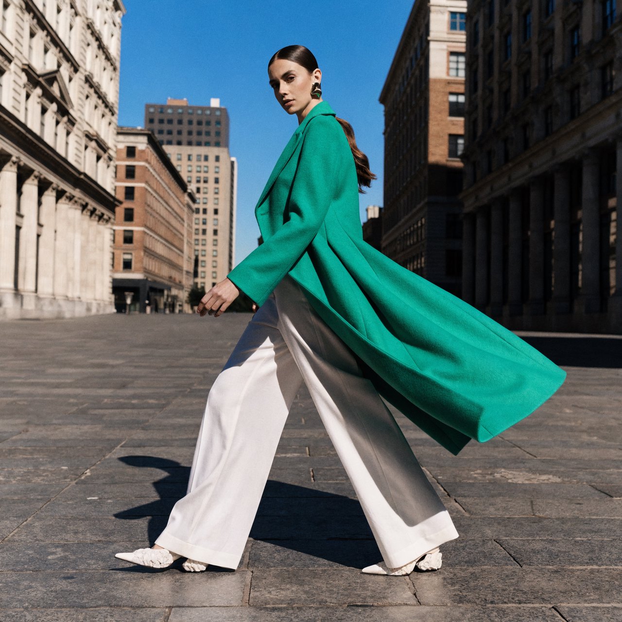 Full-length fashion editorial: model in a structured emerald wool coat and white trousers walking across a wide empty plaza, harsh midday sun, long shadow. — Recraft v4 Pro