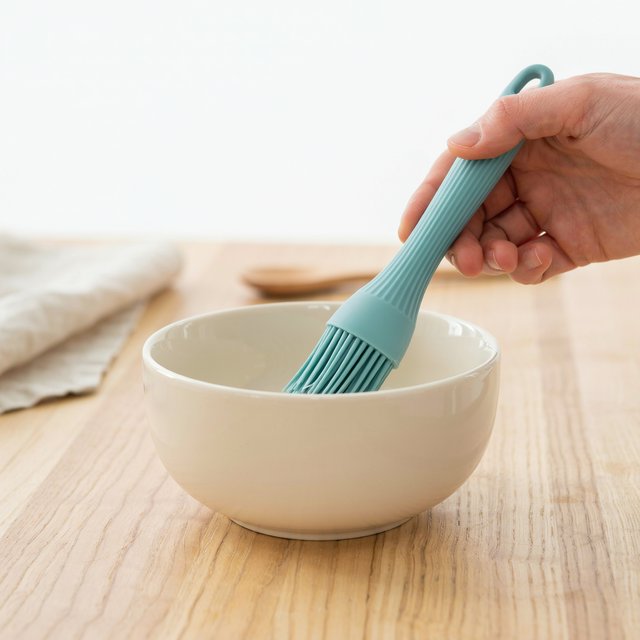 Across a honed maple counter, an adult hand guides an unbranded silicone pastry brush over a ceramic bowl; its ribbed teal handle catches diffused key light and warm bounce against a cream kitchen background, face cropped out, no lettering, commercial product image. — Ideogram v4