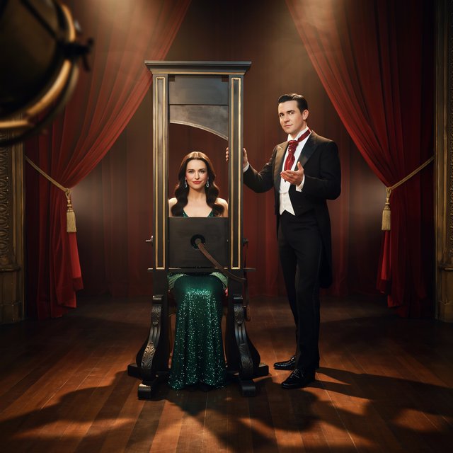 Well dressed stage magician performs the guillotine illusion using a beautiful glamorous adult female assistant. The smiling confident assistant has her head through the hole in the guillotine device. — Ideogram v4