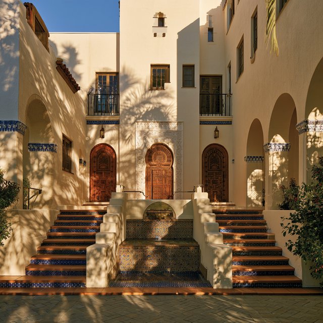 From a shaded courtyard entrance, Moorish Revival residences enclose a tiled fountain court, with cream stucco, carved cedar doors, and blue ceramic bands; late sunlight filters through arcades, revealing aligned walls and credible supports. — Midjourney V8.2