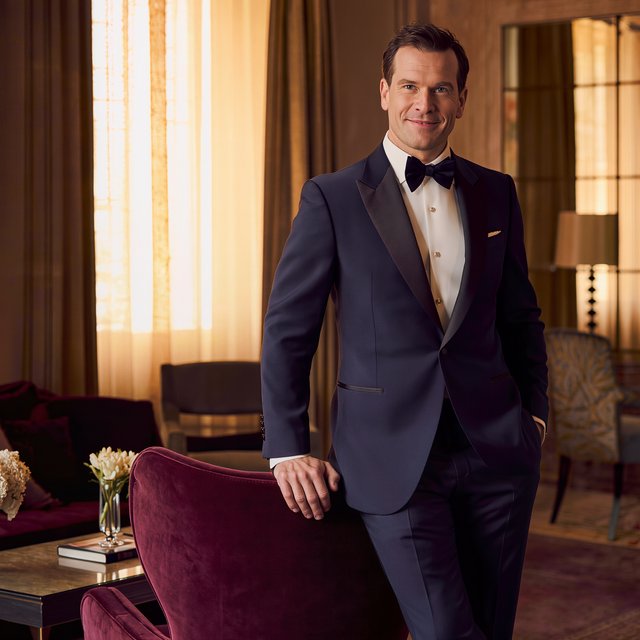 Editorial glamour photograph of an adult man in a midnight-blue tuxedo, standing naturally beside a velvet chair in a quiet hotel lounge, flattering amber window light, relaxed expression, polished magazine composition. — Kling v3