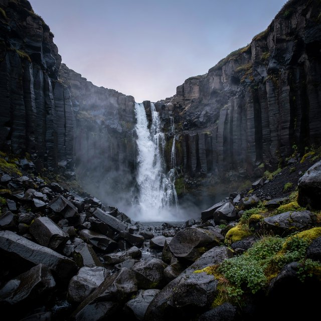 After a passing squall, a waterfall drops through a narrow basalt gorge at dusk, its white plume breaking against dark ledges; rain-beaded moss and sharp foreground stones lead toward cliffs fading behind drifting spray. — Ideogram v4.5